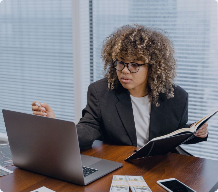 Photo of a person wearing glasses and a blazer, seated at a desk with a laptop, holding a notebook and pen while looking at the screen.